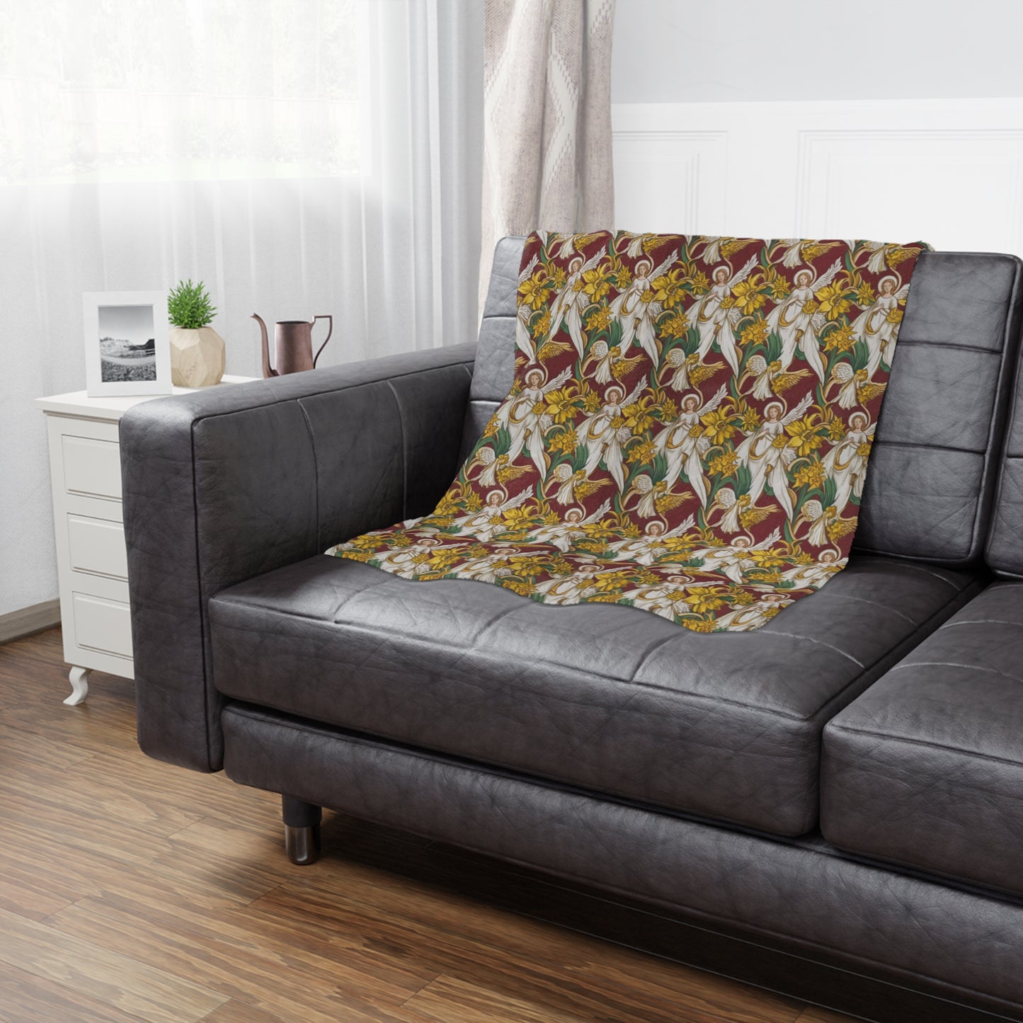 A cozy gray couch adorned with a vibrant blanket featuring angels and yellow daffodils on a maroon background. A small plant and a decorative pitcher sit on a nearby white side table, with soft natural light filtering through sheer curtains.