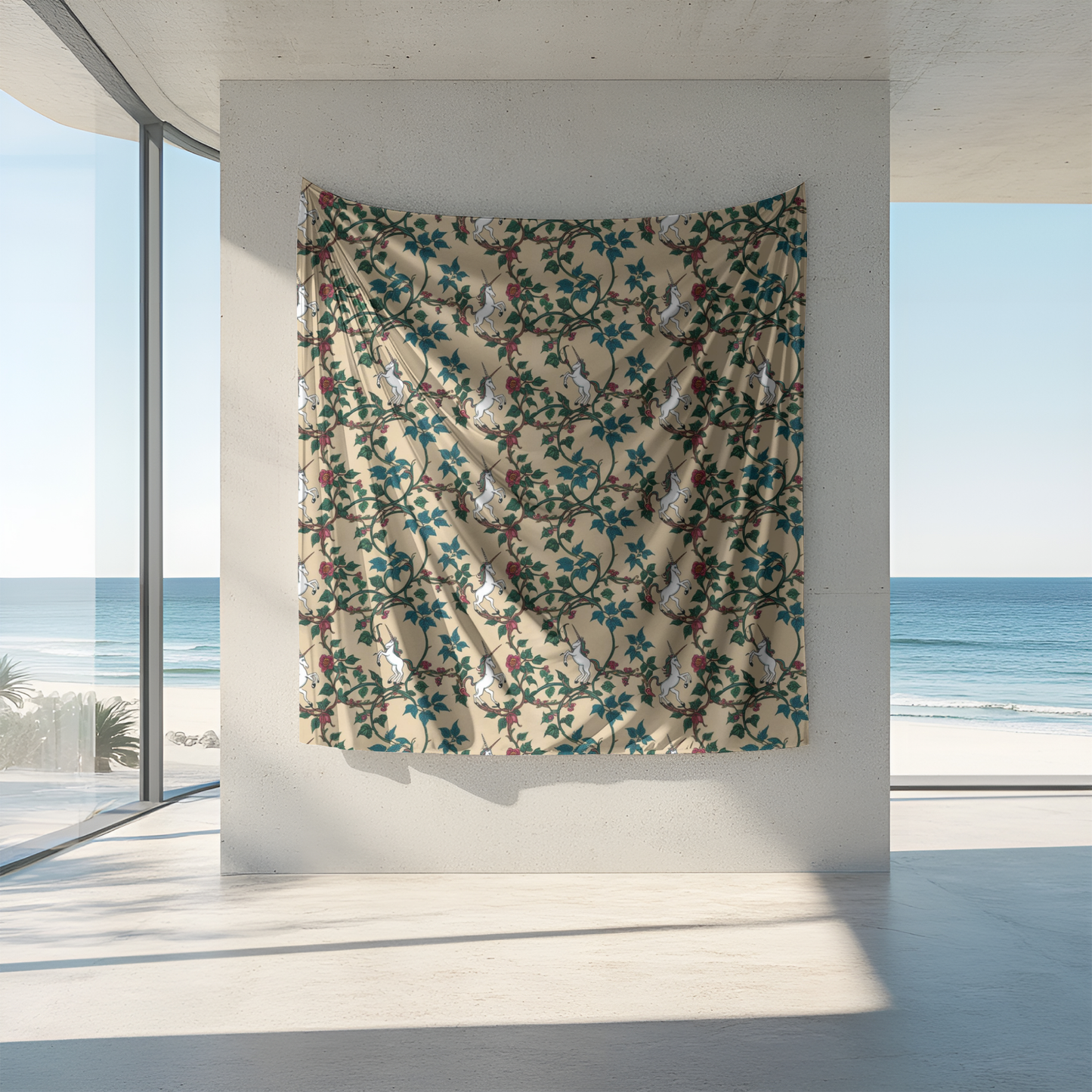 A patterned tapestry hangs on a smooth concrete wall, featuring white rabbits and floral designs in green and red against a beige background. Bright sunlight streams through large windows, illuminating the serene beach view beyond.