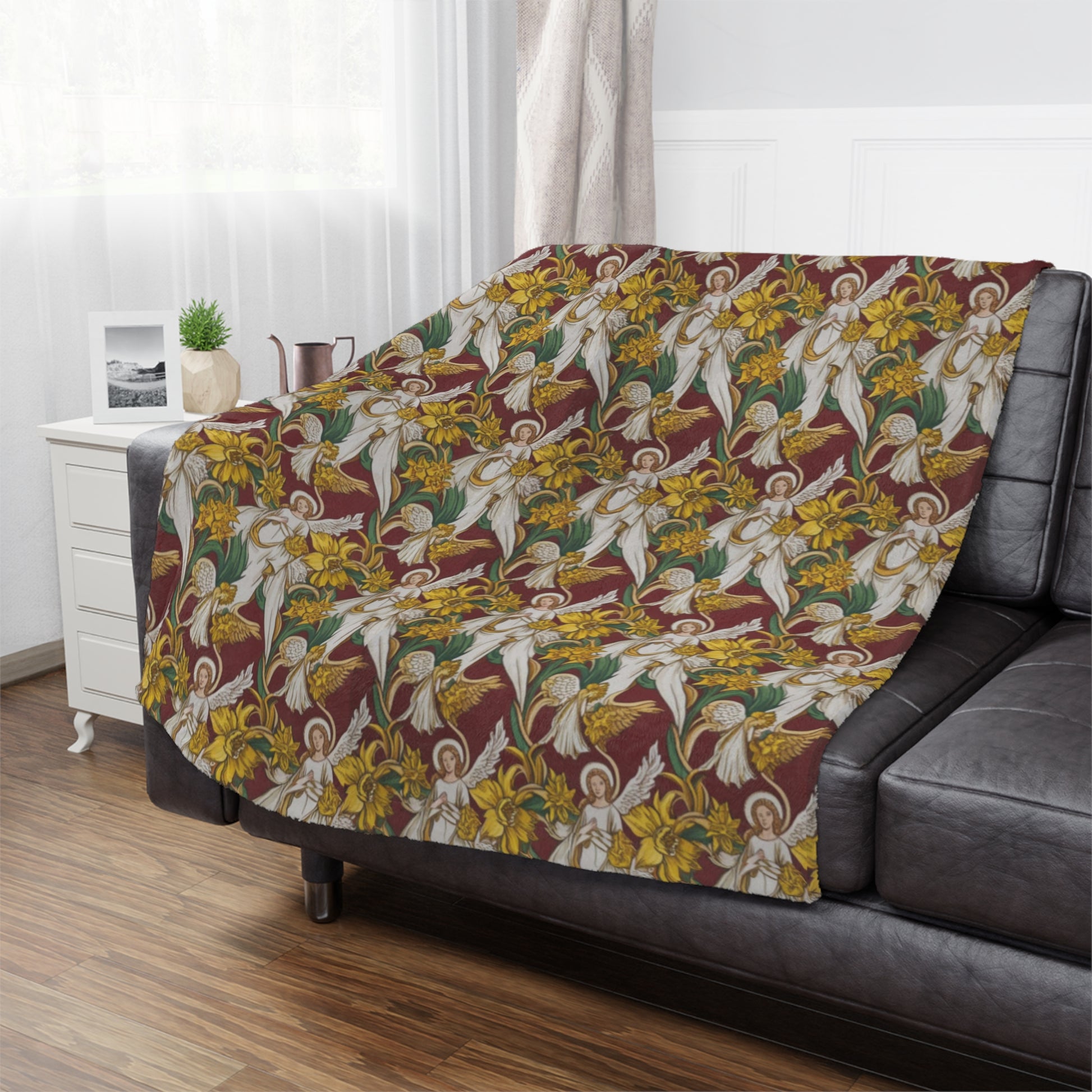 A cozy gray couch adorned with a vibrant blanket featuring angels and yellow daffodils on a maroon background. A small plant and a decorative pitcher sit on a nearby white side table, with soft natural light filtering through sheer curtains.
