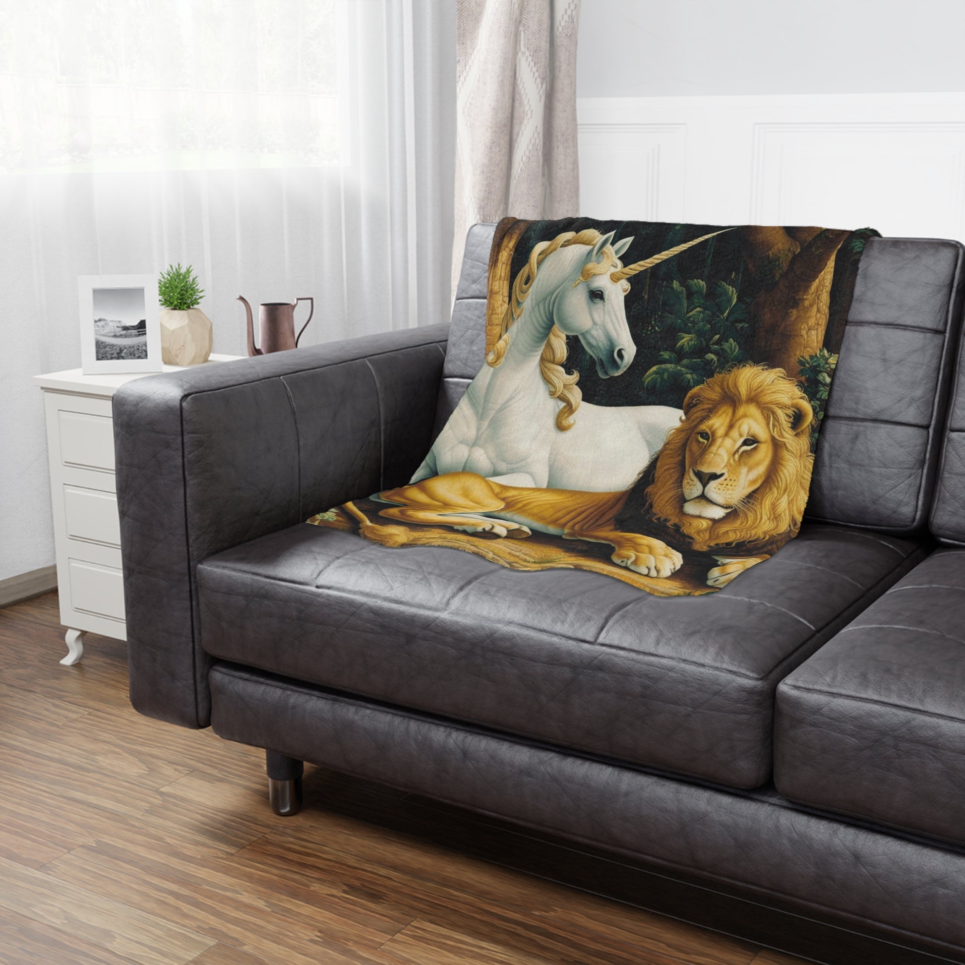 A cozy black leather couch features a blanket depicting a white unicorn and a golden lion resting together. A small plant and a decorative pitcher sit on a nearby white side table, with soft natural light streaming through the window.
