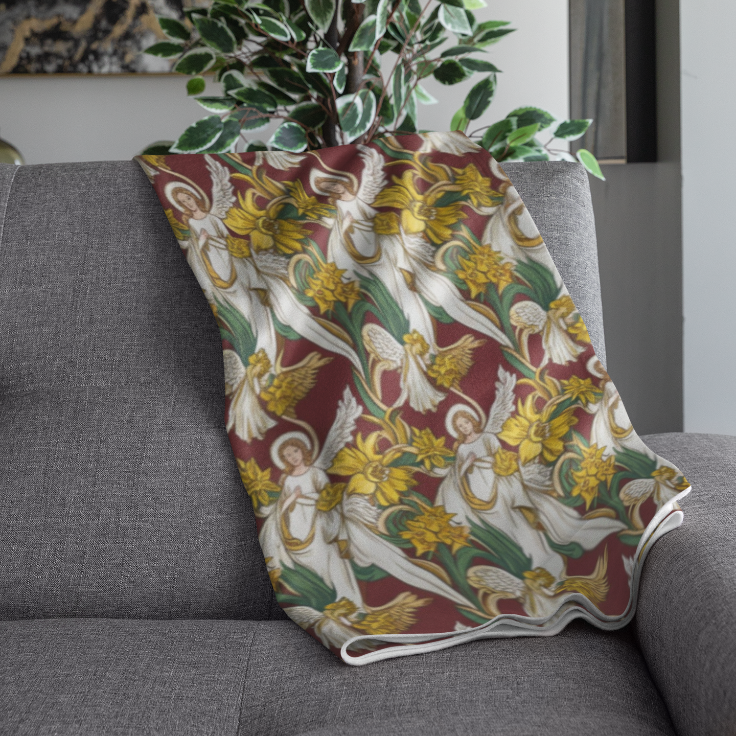 A decorative blanket draped over a gray couch, featuring a vibrant pattern of angels and yellow daffodils against a rich maroon background. A leafy plant is visible in the background, adding a touch of greenery to the scene.
